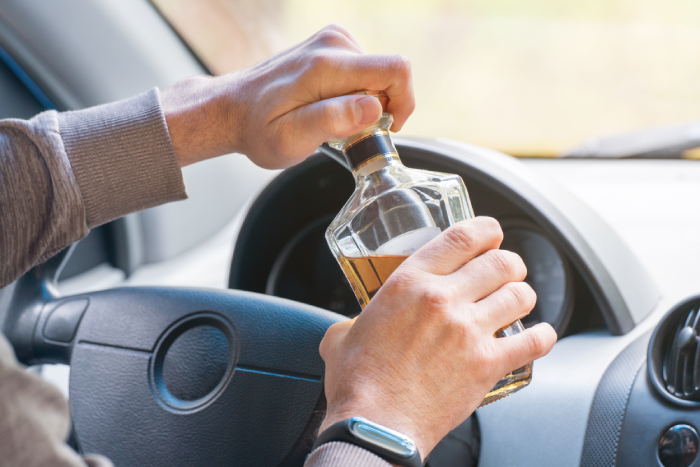 The driver is sitting behind the wheel and holding a bottle of alcoholic drink. Breaking the law and drinking alcohol while driving.