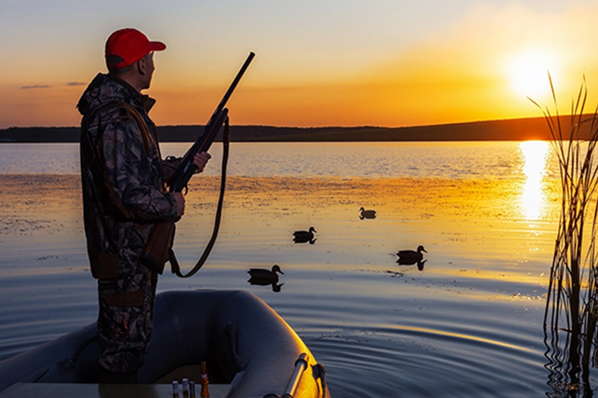 A man wearing a camouflage jacket and red cap holding a shotgun and looking towards a vibrant sunset over the water.